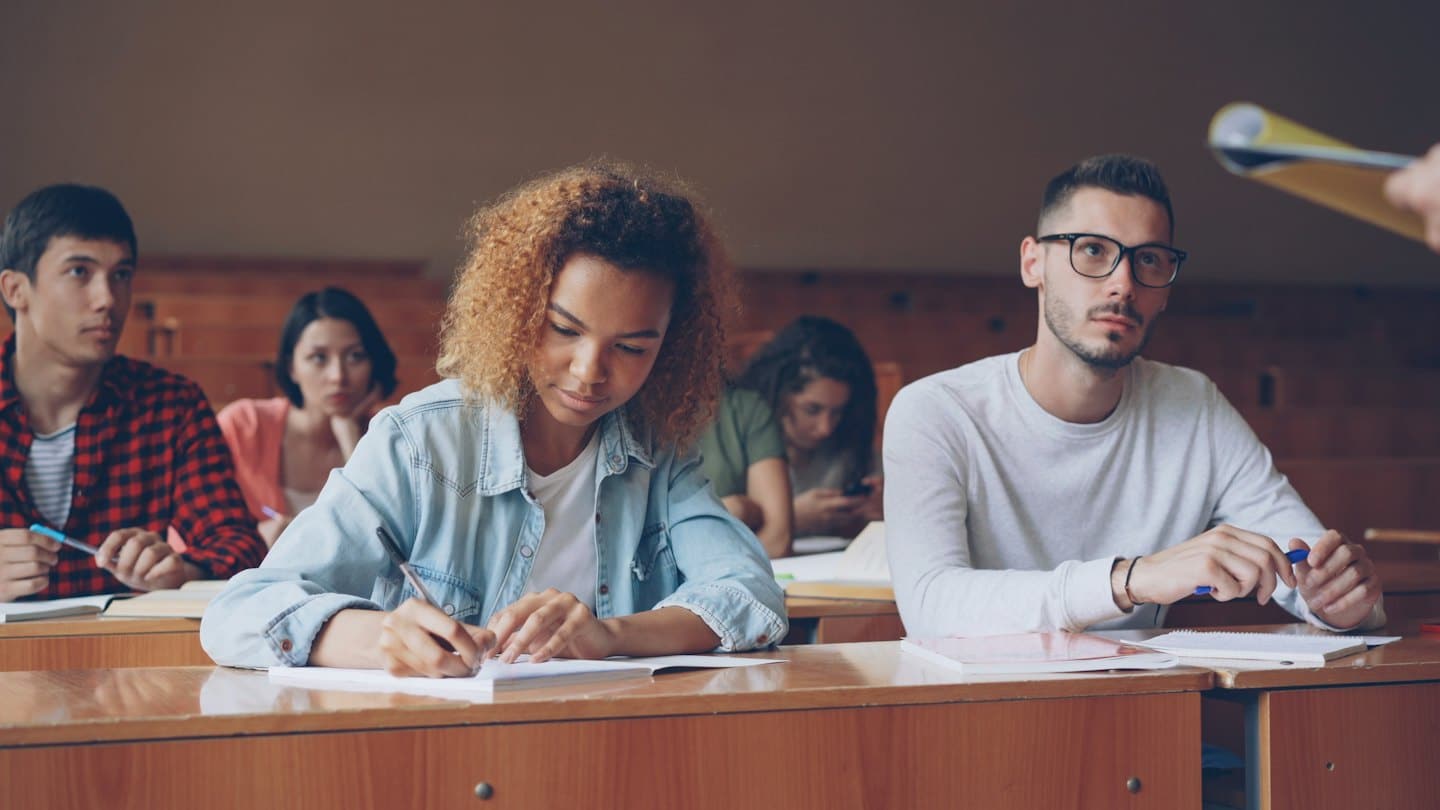 Studenti universitari ascoltano una lezione in un'aula magna in legno.